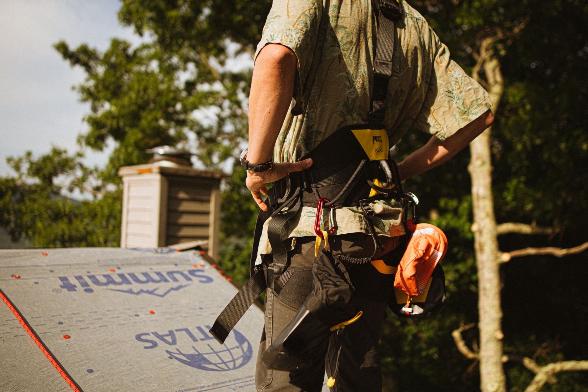 Roofer wearing a safety harness working on a rooftop under a clear sky.